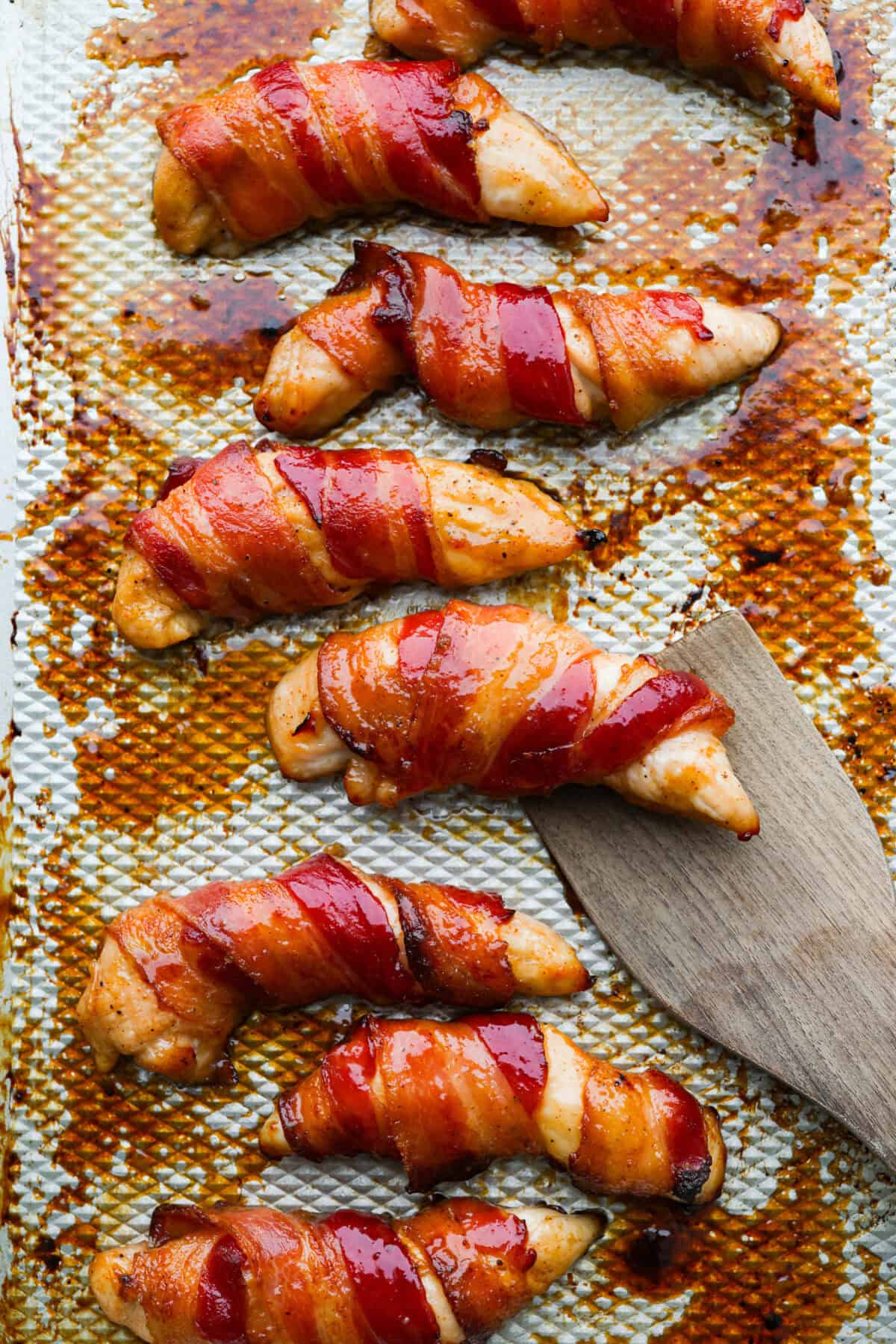 Bacon brown sugar chicken tenders are sweet, smoky, and slightly spicy. Made with only a few simple ingredients, they're like candied bacon hugging juicy chicken without being overly sweet. Great as a main or appetizer, and always a total hit! Bacon brown sugar chicken tenders cooked and on a baking rack with a wooden spatula.