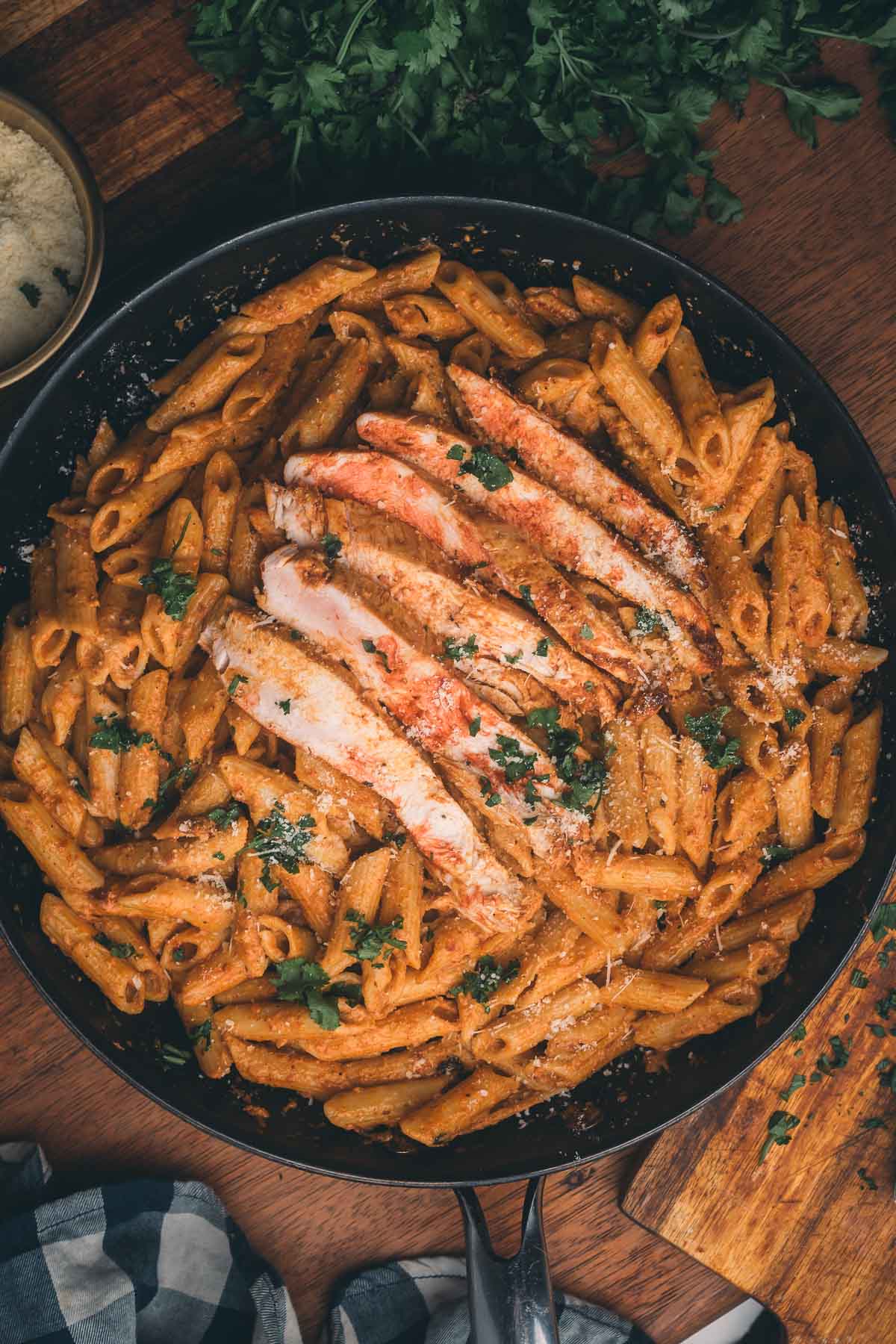 A skillet filled with penne pasta in a creamy sauce, topped with sliced grilled chicken and garnished with chopped parsley.