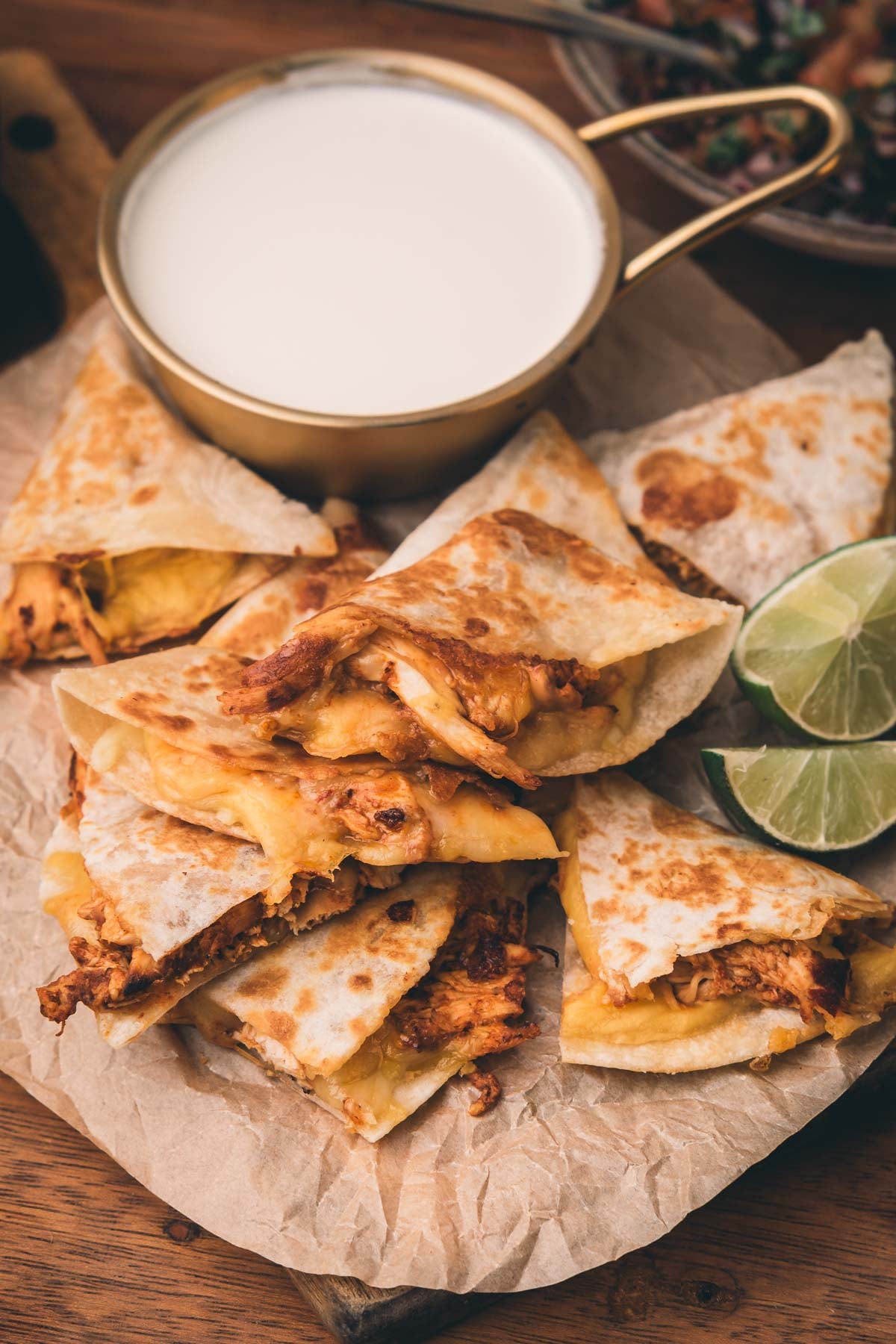 Chicken quesadilla wedges arranged on parchment paper with a cup of white dipping sauce and lime slices on the side.