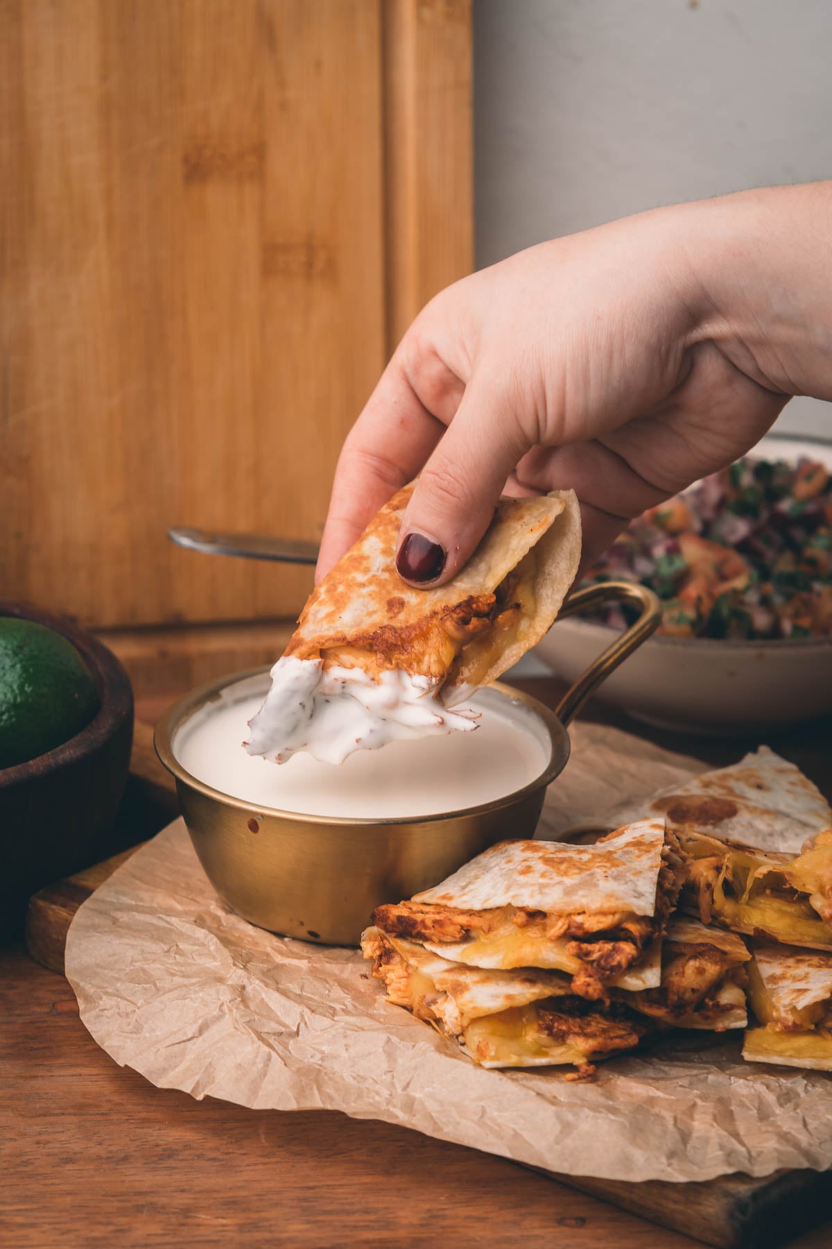 A hand dips a piece of quesadilla into a bowl of white sauce, with more quesadilla slices and a bowl of salsa nearby on a wooden surface.