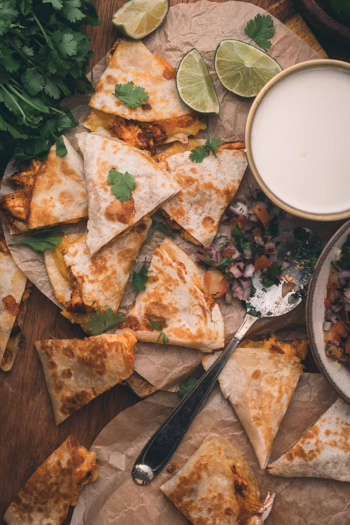Sliced quesadillas on parchment paper with lime wedges, fresh cilantro, a bowl of sour cream, and a spoonful of salsa on a wooden table.