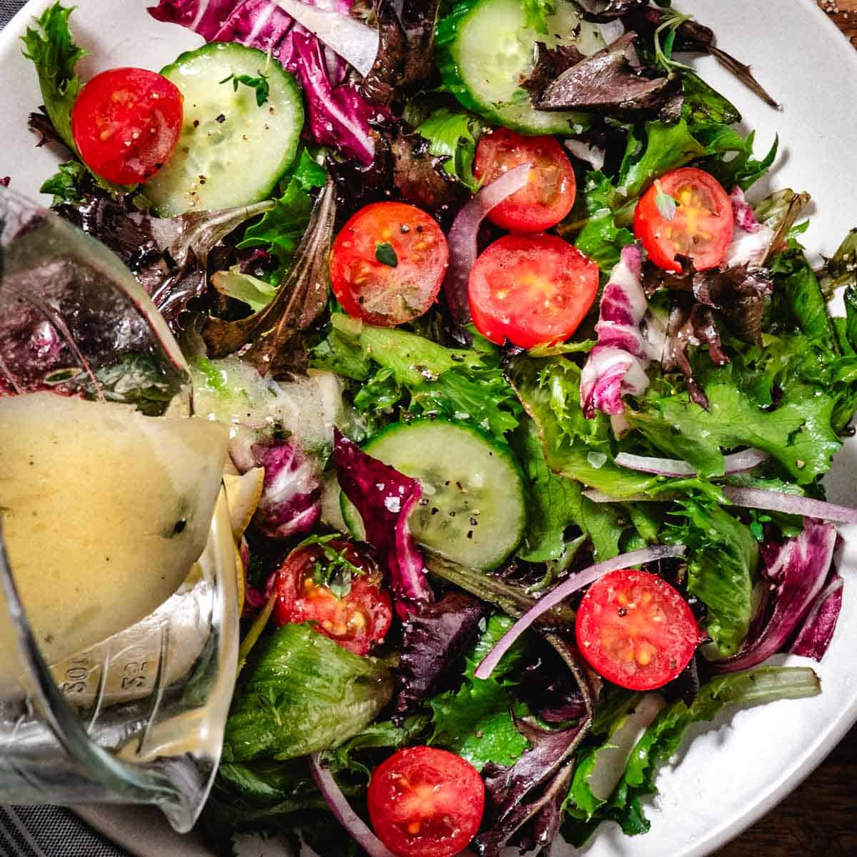 Mixed garden salad with cucumbers, cherry tomatoes, and red onion as lemon Dijon dressing is poured over fresh greens