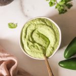 Avocado cilantro sauce in a white bowl with a spoon resting on the side.