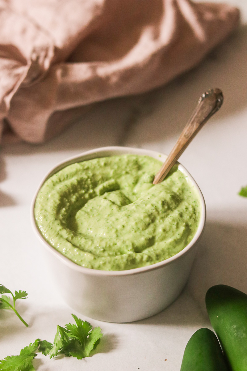 a small white bowl filled with avocado cilantro sauce with a spoon resting on the side.