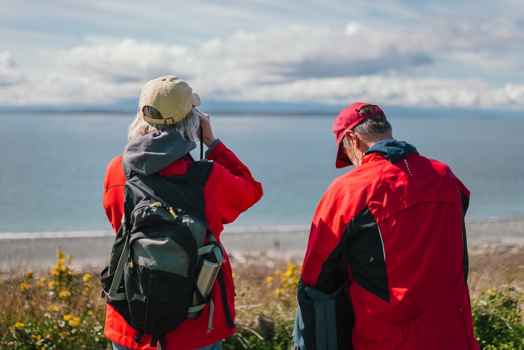 Elderly couple in red jackets enjoying seaside view, outdoors with backpacks and travelling more after retirement