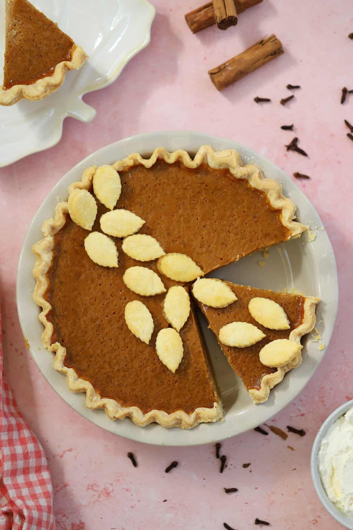 A gluten free pumpkin pie on a pink backdrop with cinnamon sticks and cloves surrounding it.