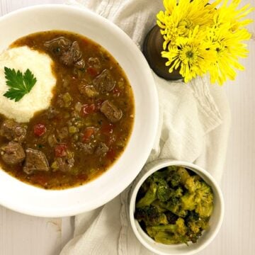 Grillades and grits on a white marbled surface with a napkin, flowers, and broccoli side dish.