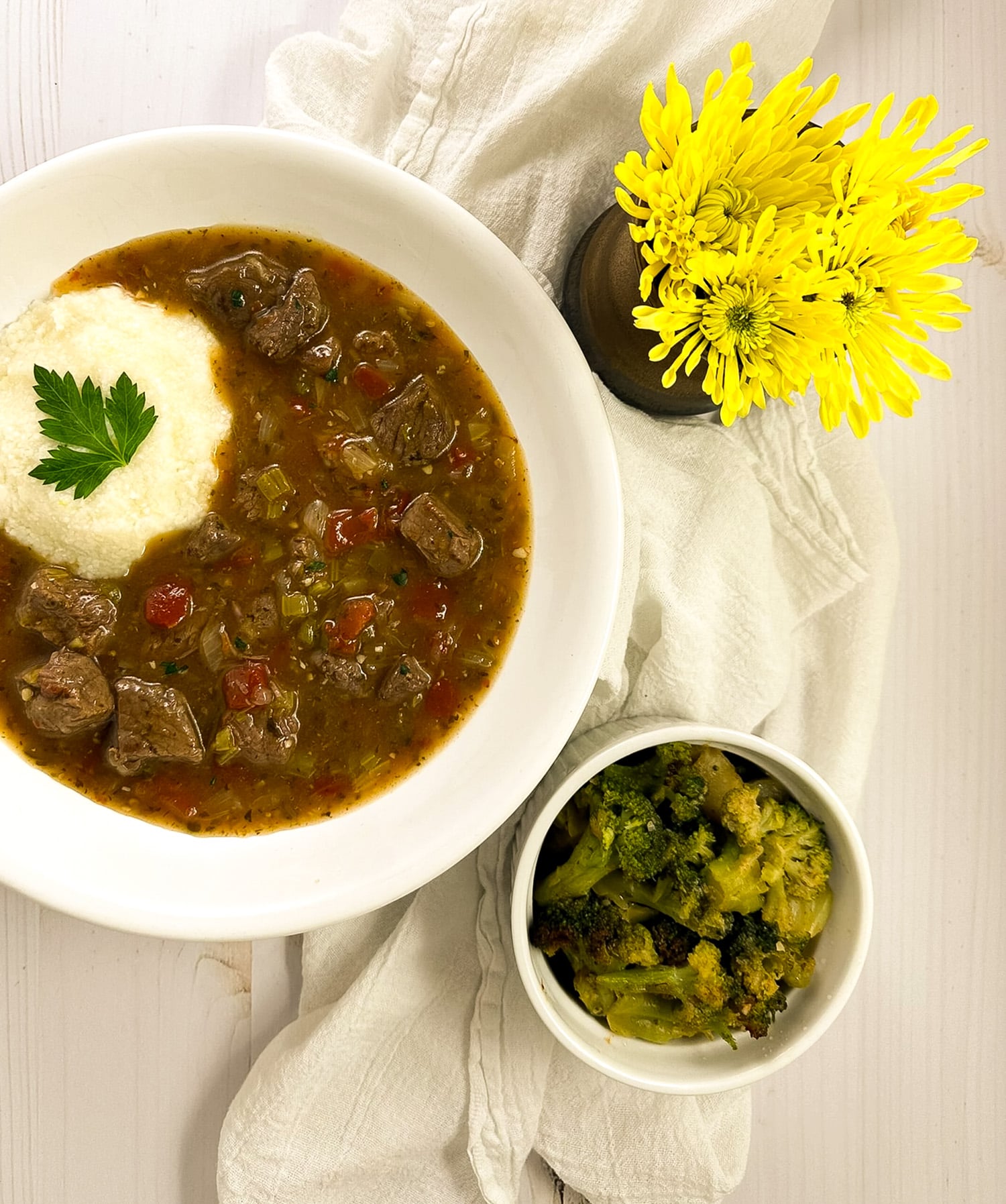 Grillades and grits in a round white bowl alongside a white napkin, yellow flowers, and a small bowl of broccoli.
