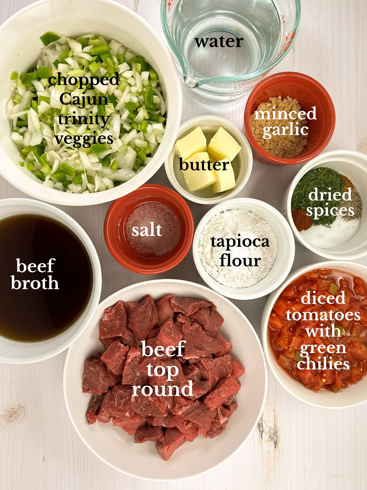 Labeled ingredients for grillades and grits in bowls on a white marbled background.