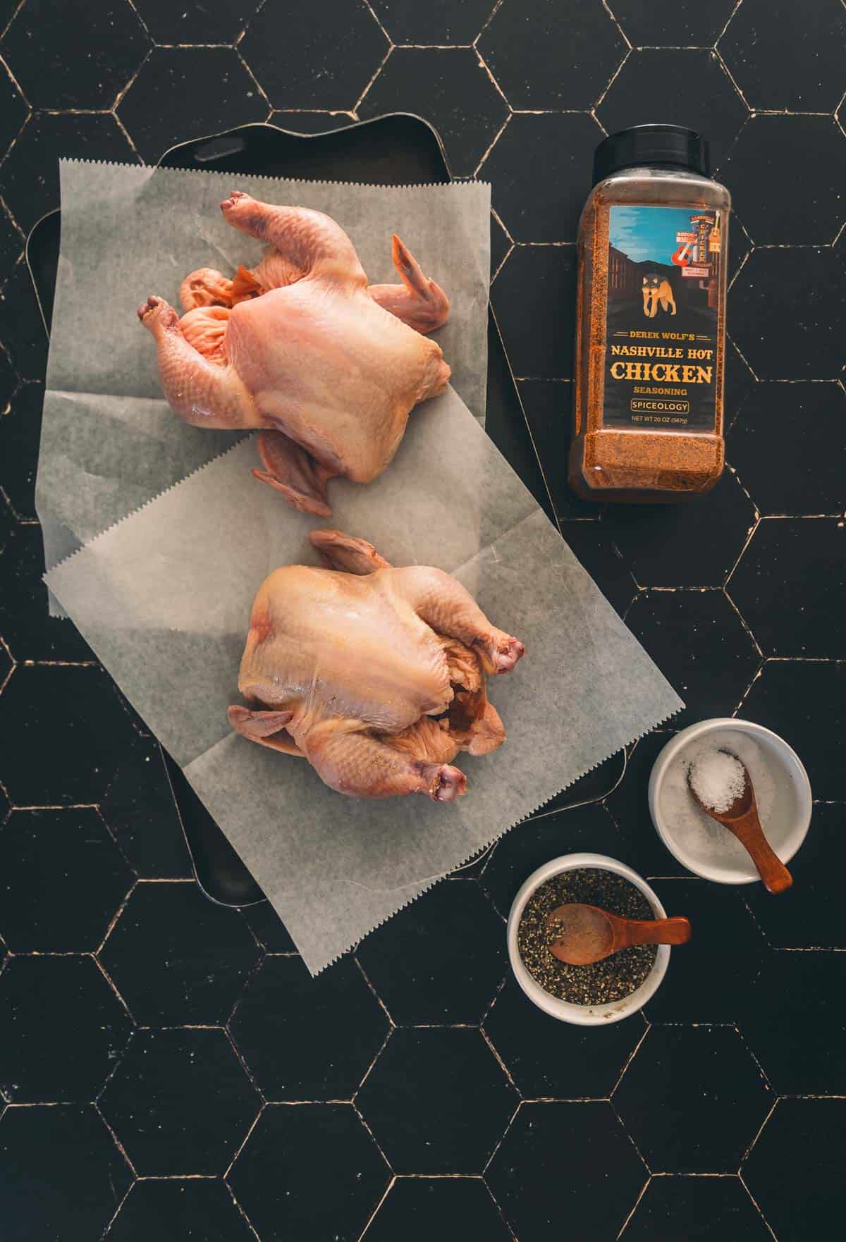 Two Cornish hens on a baking sheet next to a jar of seasoning.