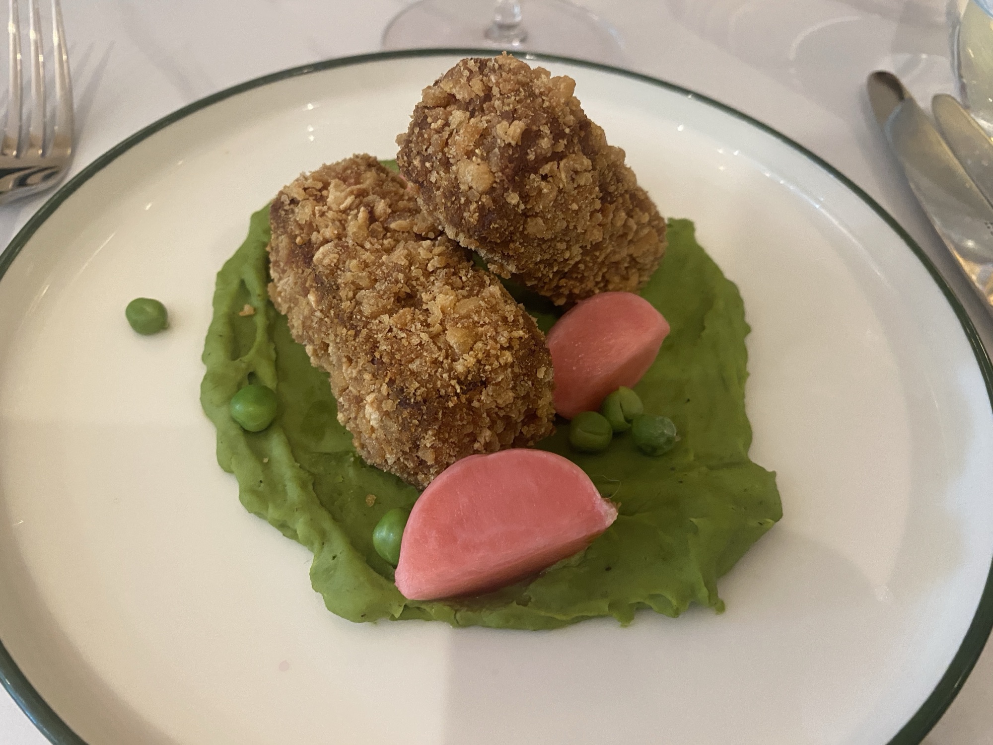 A white plate with a green rim, sitting on a white table cloth, with a knife and fork either side. In the middle the plate are two crispy, breaded croquettes of ham hock terrine, sitting on a minted pea puree, with a few scattered peas and a couple of quarters of pink pickled radish.