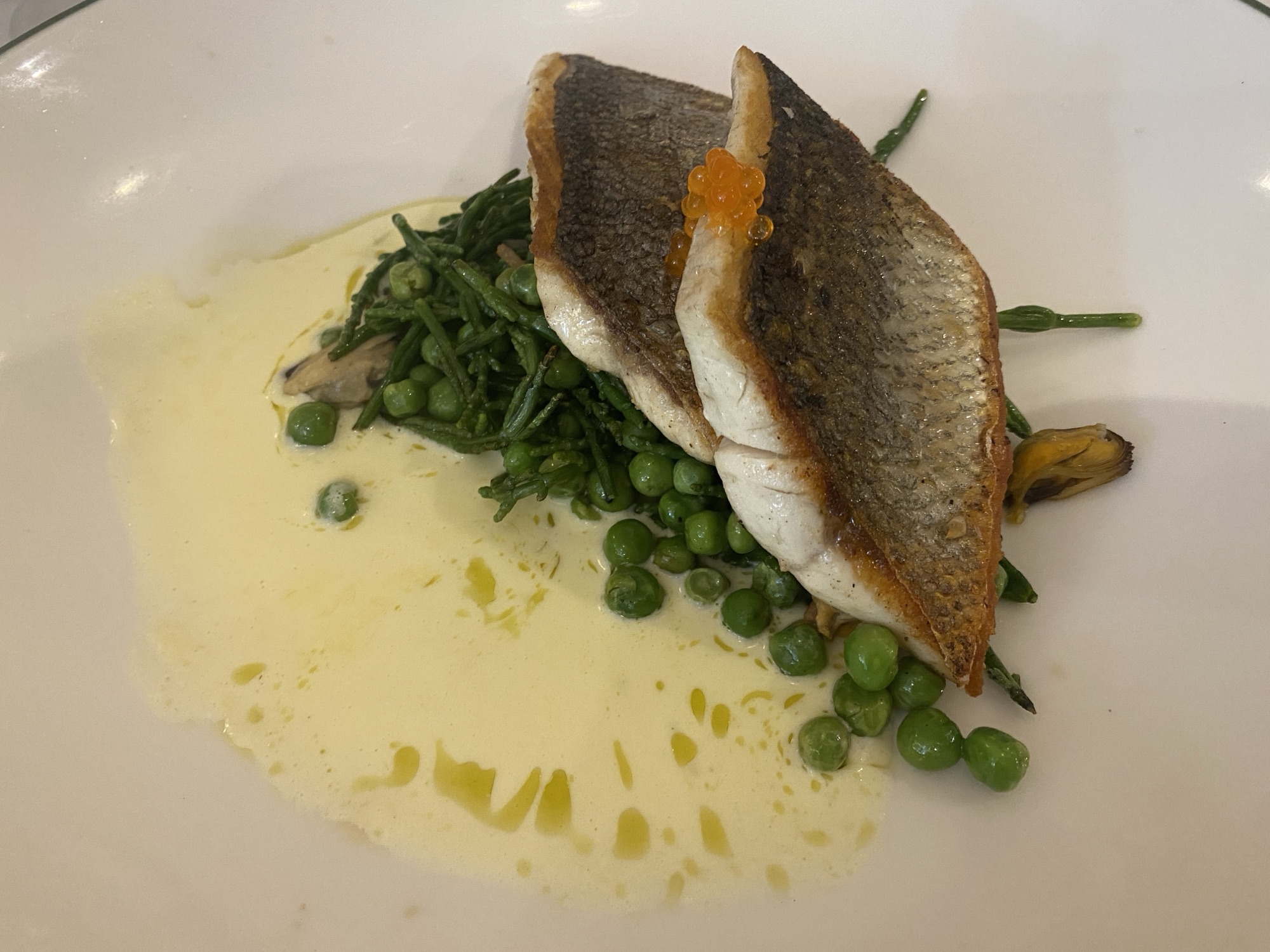 A close up of a white plate. Two crispy skin-on white fillets of seabass are nestled on top of a pile of peas and samphire. All of which sits in a puddle of creamy sauce.