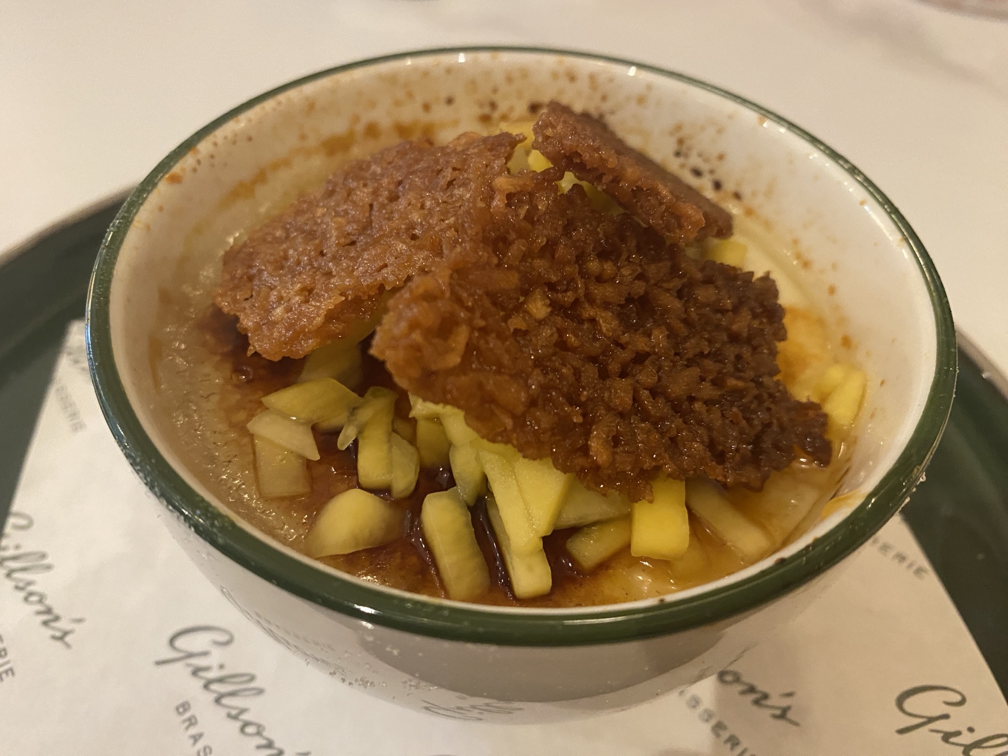 A close up of a white bowl of creme brulee with a green rim. It's almost completely covered in slivers of mango, and on top of that are arranged three pieces of crunchy, sugary, oaty biscuit.