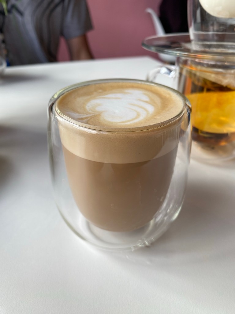 This is a decaf flat white coffee in a double walled glass mug, sitting on a white table with a tea in the background 