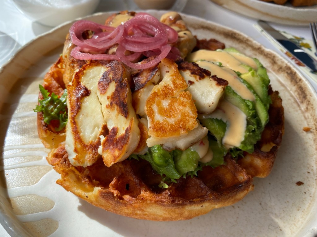 Loaded gluten free round waffle on a ceramic plate. Piled up with halloumi, avocado, salad leaves, pink pickled onions and a drizzle of pale sauce