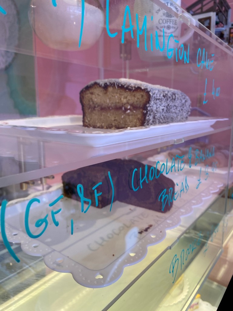 Glass cake counter with blue writing on the front. Inside is a gluten free Lamington cake at the top (the writing says so) and a gluten free chocolate banana bread