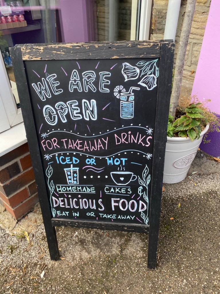 Wooden A frame sign advertising the cafe is open for drinks, cakes and food, to eat in or take away 