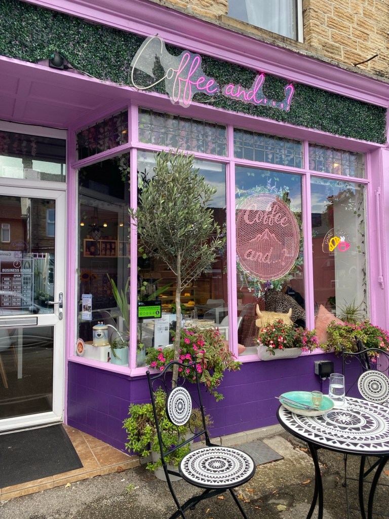 Picture of the outside of Coffee and...? on Abbeydale Road. It's got pale pink paint and lots of greenery, with a big crocheted sign int he window. There is a set of mosaic table and chairs outside