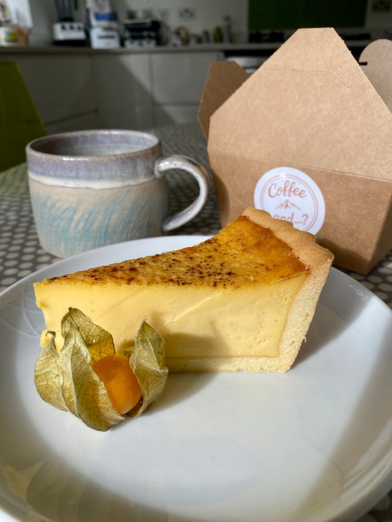 White plate with a rather lovely looking slice of gluten free egg custard, accompanied by a single, decorative physalis. In the background sit a hand crafted ceramic mug and an open, brown, cardboard cake box