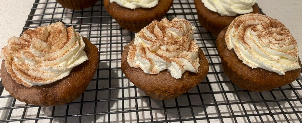 A cooling rack with three brown coffee cupcakes with a whipped cream and mascarpone topping, sprinkled with chocolate powder. Others are in view at the back