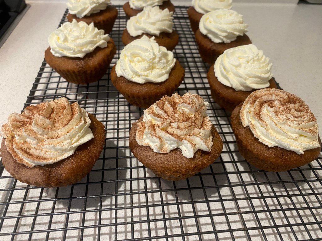 A cooling rack with brown coffee cupcakes with a whipped cream and mascarpone topping. Three at the front have been sprinkled with chocolate powder.