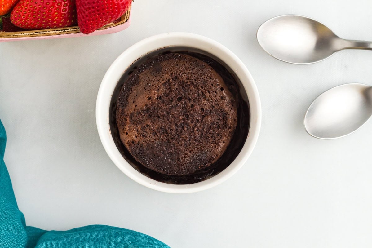 Flourless mug cake in a white ramekin next to fresh strawberries and two spoons