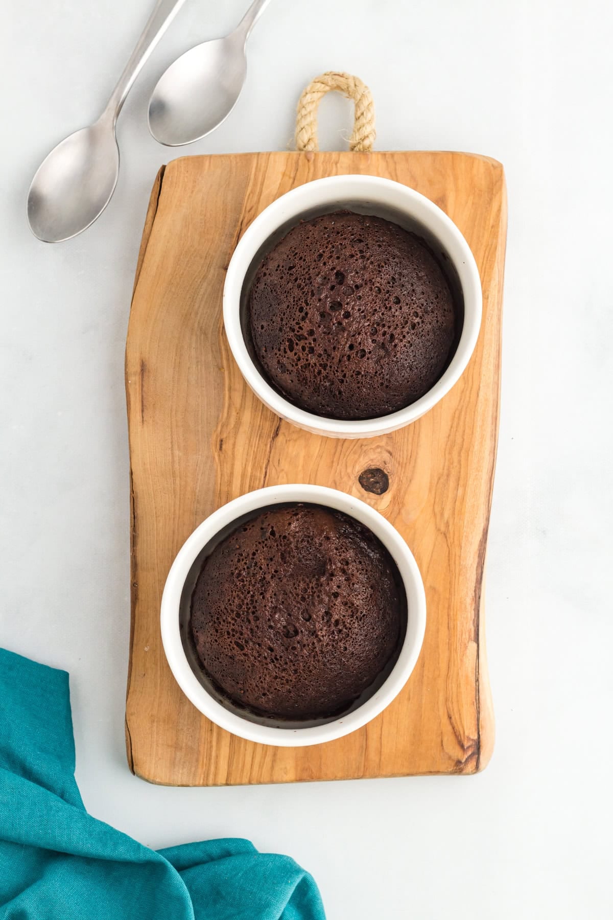Two flourless mug cakes baked in white ramekins on a wooden board with spoons and a teal napkin