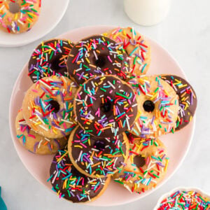 Gluten-free vanilla and chocolate cake donuts topped with rainbow sprinkles on a pink plate beside a bottle of milk.
