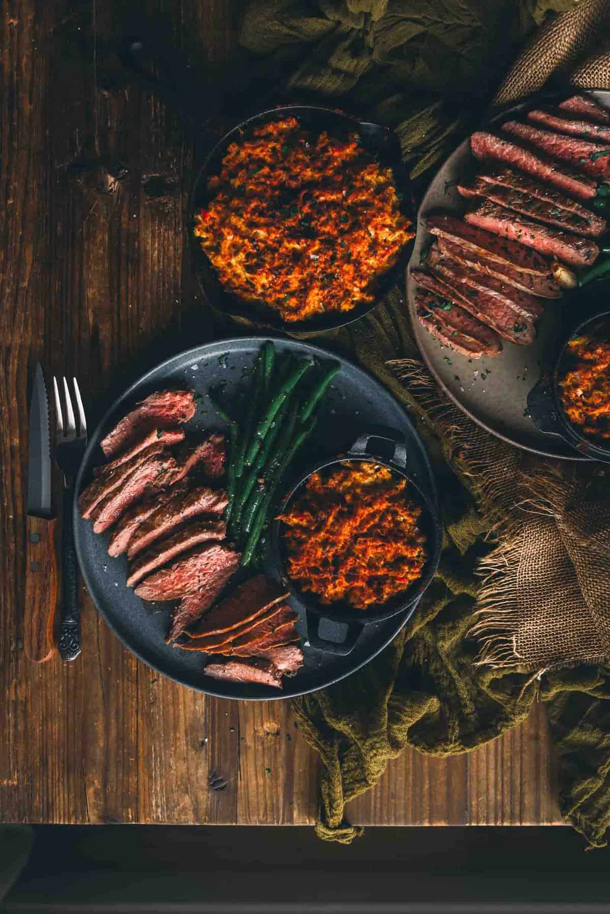 Overhead rustic table shot of sliced cooked cast iron steaks and side dishes.