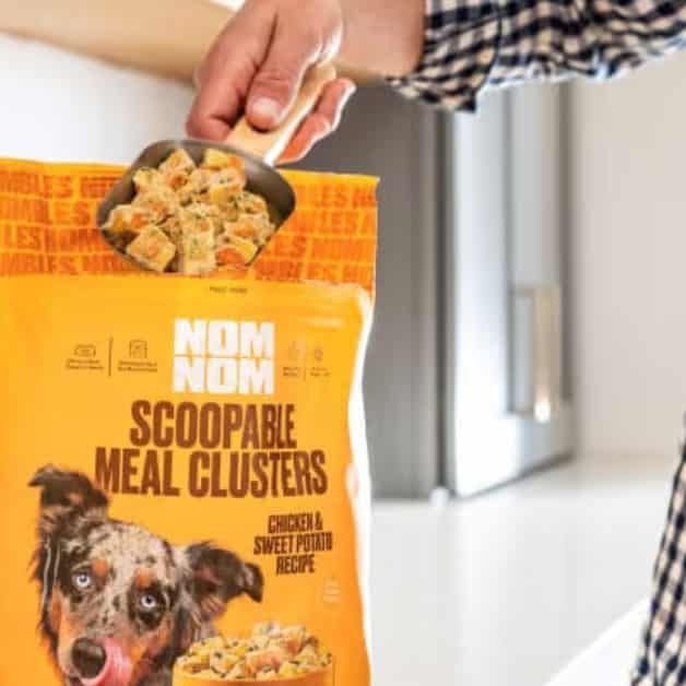 Hand holding a scoop of chicken and sweet potato clusters above an open Nom Nom Scoopable Meal Clusters bag