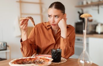 Upset woman looking at delicious piece of high-calorie pizza, holding slice in hand. Diet and digestive problems as well as spoiled and tasteless food