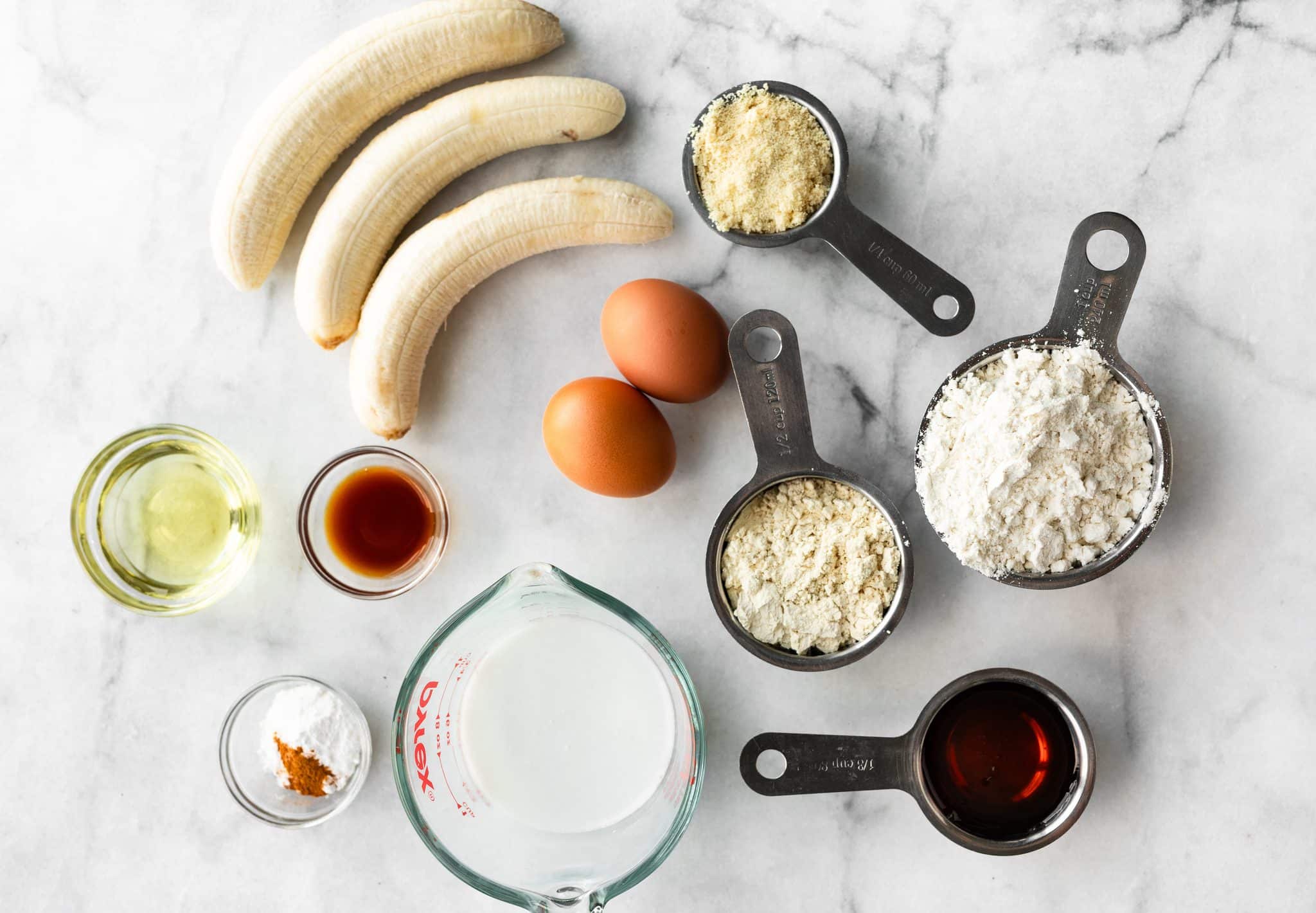 Bananas, eggs, flour, protein powder, milk, oil, maple syrup, vanilla, and spices arranged on a marble surface.